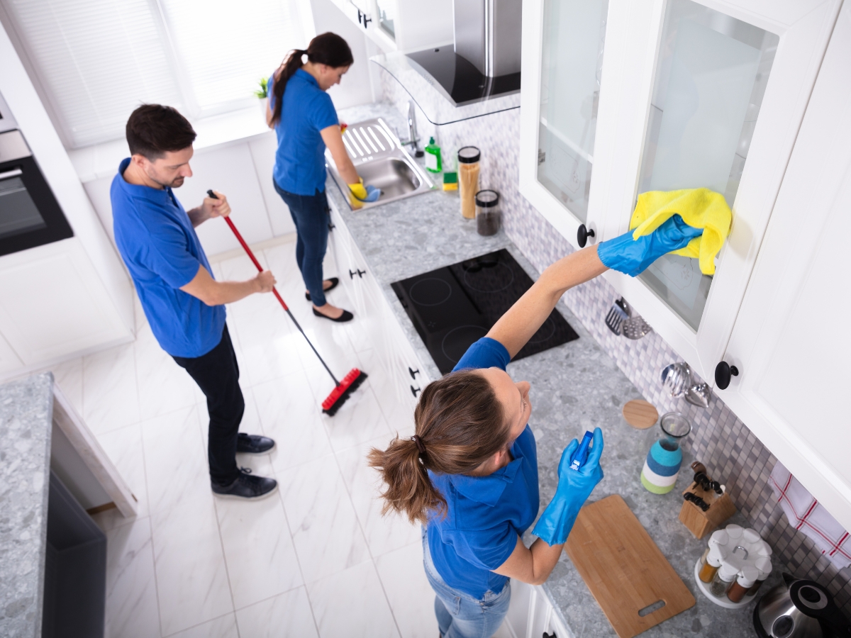 Three people in blue shirts clean a modern kitchen, showcasing Holiday Home Maintenance Tips: one wipes a cabinet, another sweeps the floor, and the third washes dishes at the sink.