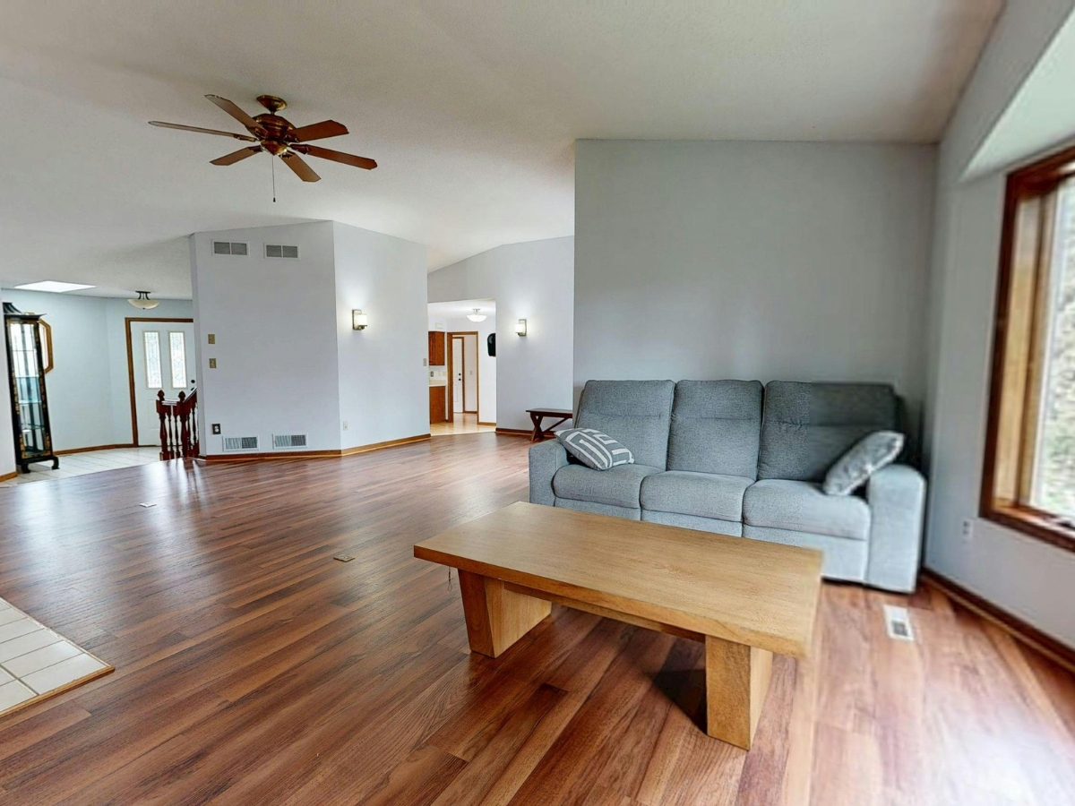 Spacious living room with hardwood flooring offering added benefits, gray sofa, wooden coffee table, large window, and ceiling fan; open layout connects to other rooms.