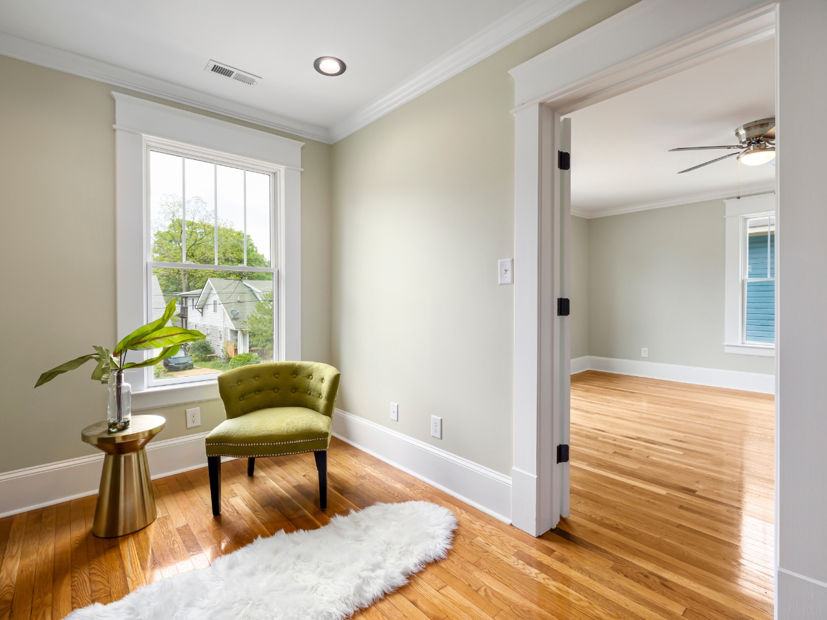 A small seating area with a green chair, side table, and white rug sits by a window, leading to an empty adjoining room with wood laminate floors—perfect for considering laminate pros and cons in your home.