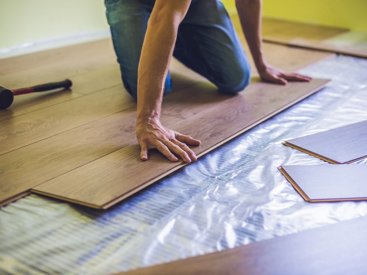 A person kneels on the floor installing wooden laminate flooring, using their hands to align the planks over a plastic sheet—a process often weighed by homeowners considering laminate pros cons.