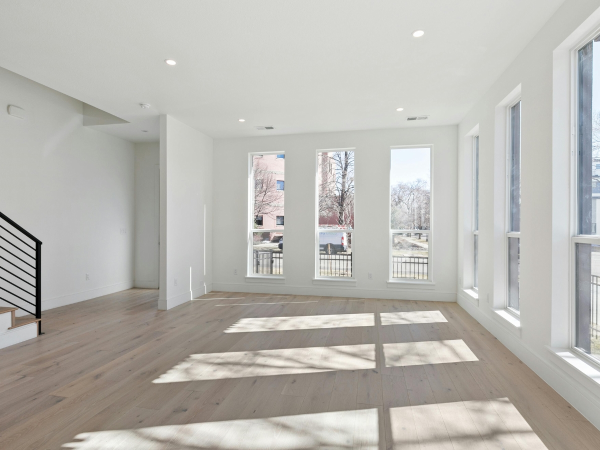 Bright, empty room with large windows, light wood flooring—explore laminate pros and cons in this airy space with white walls and a black-railed staircase on the left.