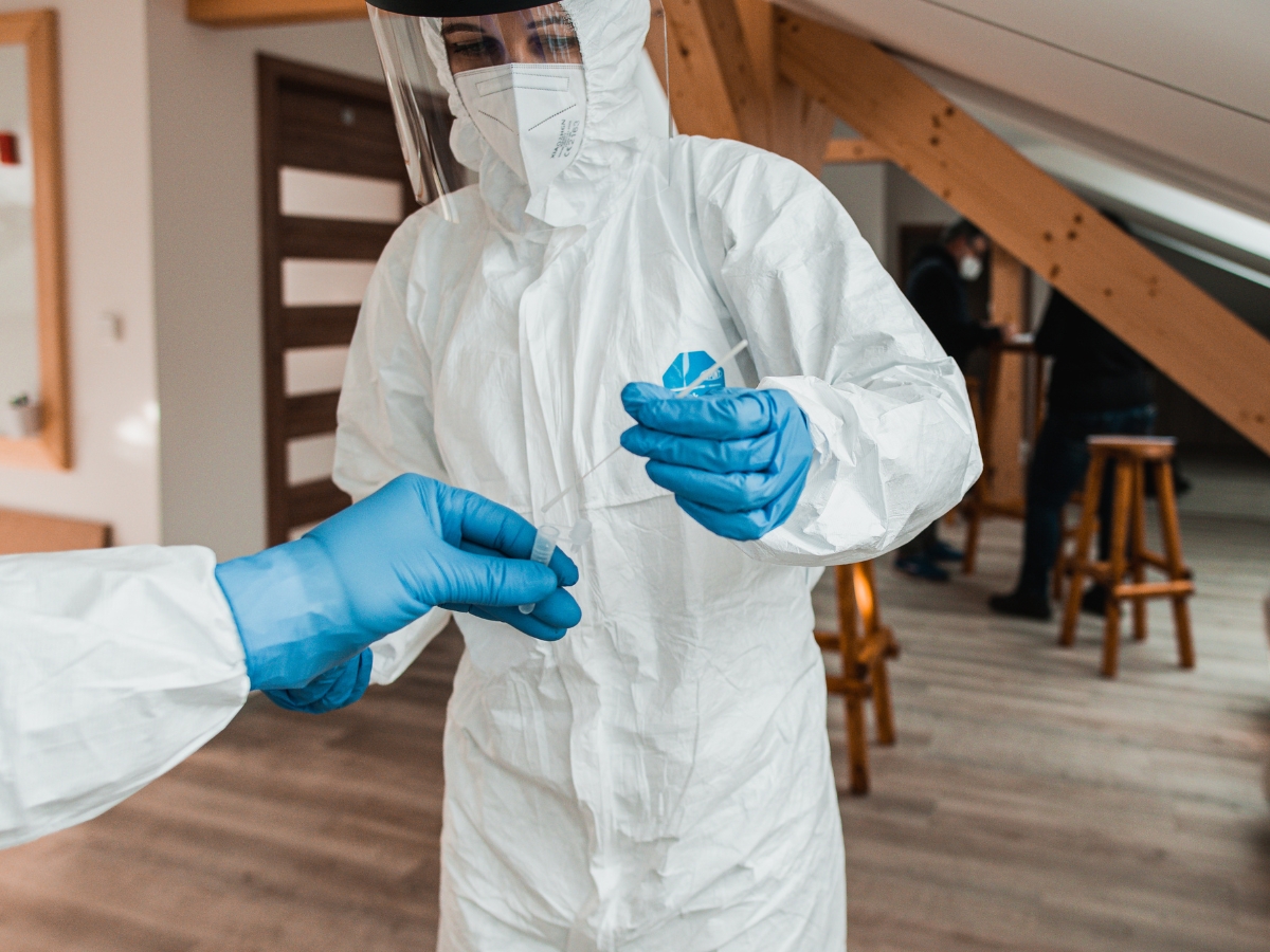Two people in full protective suits and gloves handle a swab in a room with wooden beams and stools, demonstrating asbestos awareness as one passes the swab to the other.