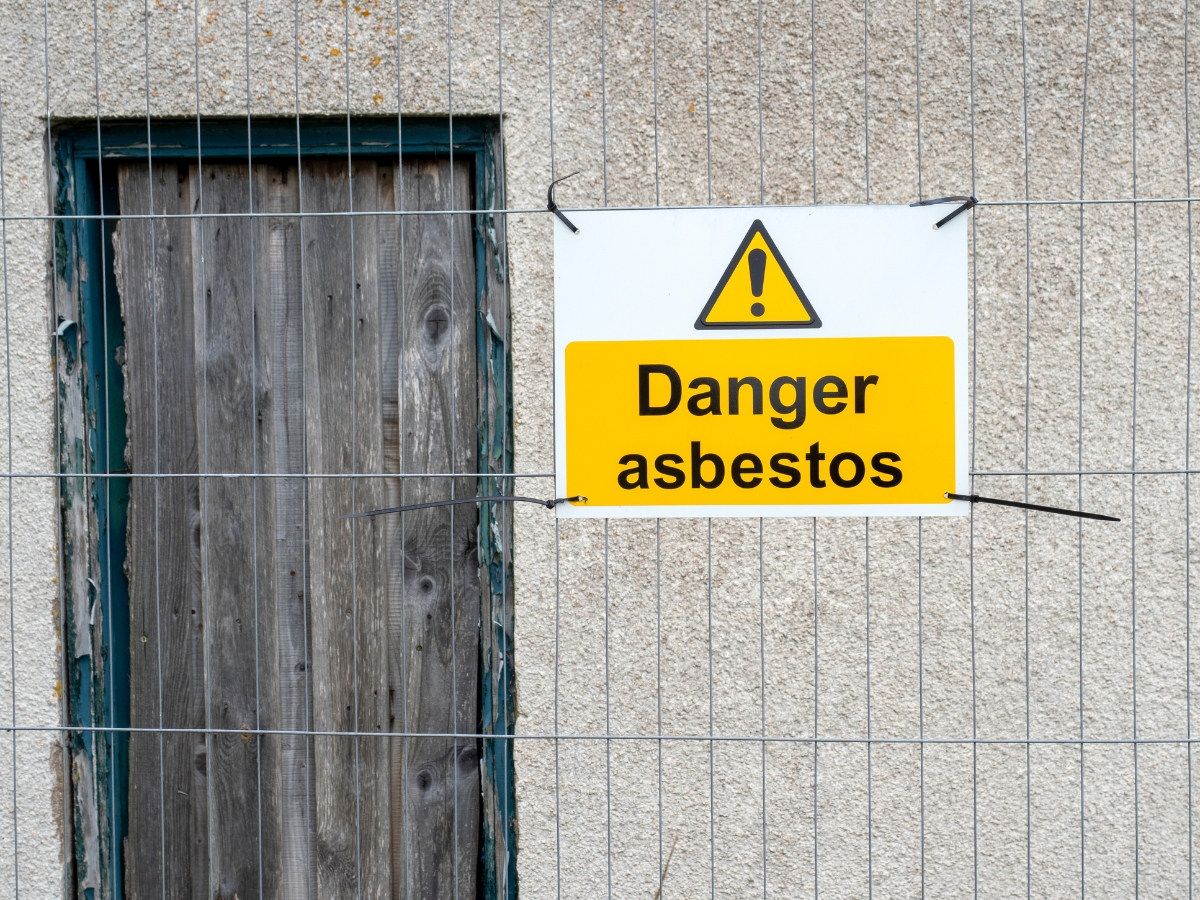A warning sign on a wire fence reads "Danger asbestos," highlighting the importance of asbestos awareness, next to a sealed wooden door on a concrete wall.