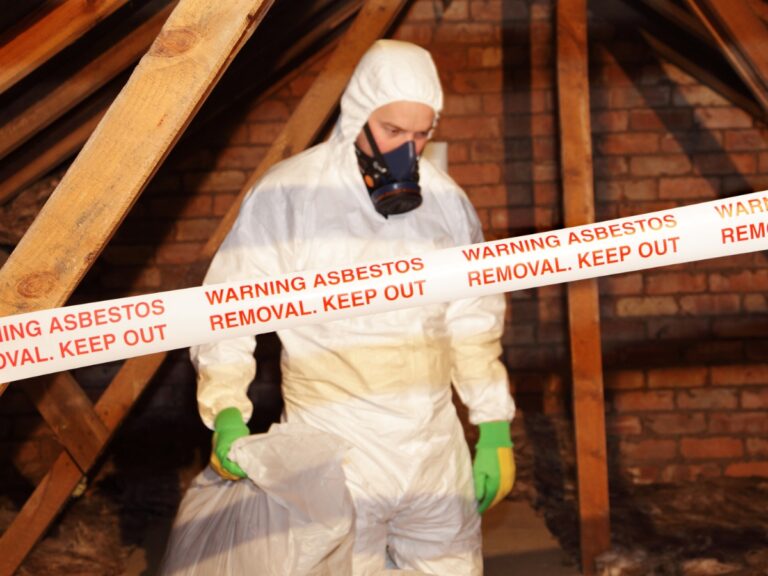 Person in protective suit and mask handles a bag behind tape reading "Warning Asbestos Removal. Keep Out," promoting asbestos awareness in an attic with exposed beams and brick walls.