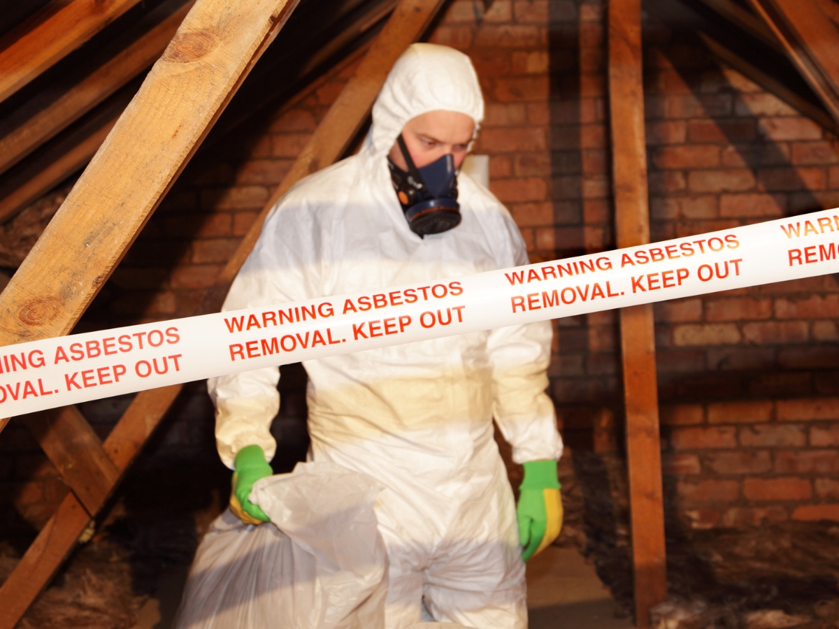 Person in protective suit and mask handles a bag behind tape reading "Warning Asbestos Removal. Keep Out," promoting asbestos awareness in an attic with exposed beams and brick walls.
