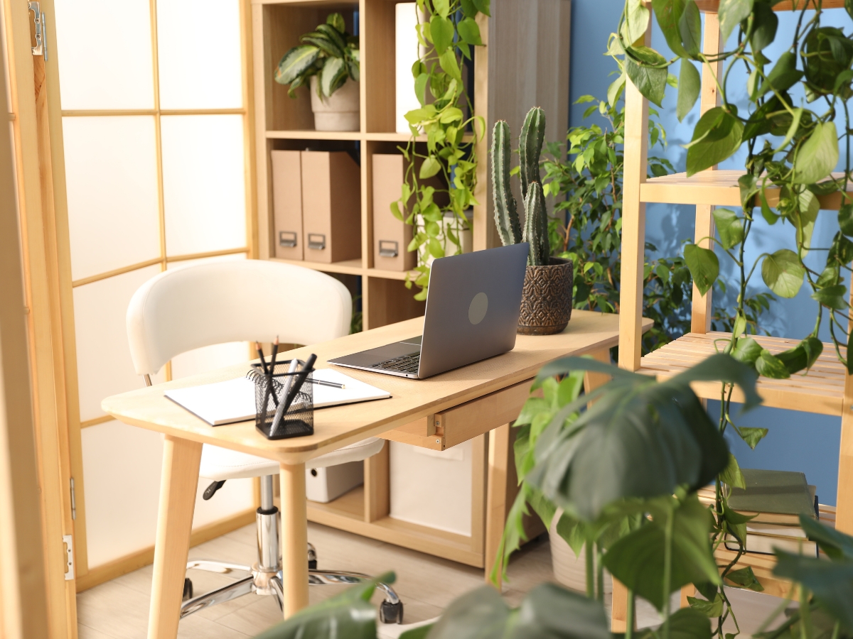 A home office with a laptop on a wooden desk, a white chair, notebooks, pens, and various green plants embraces biophilic design to create a refreshing workspace.