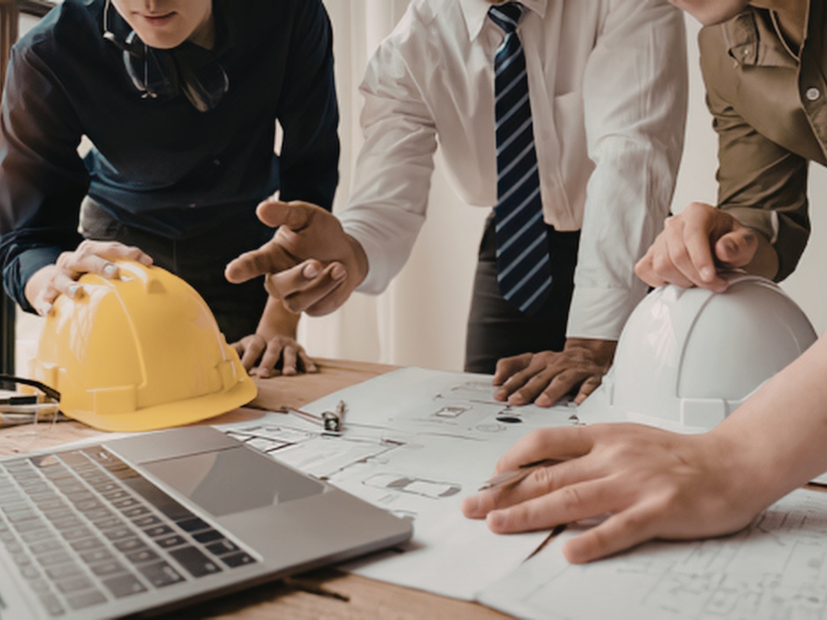 Three people in business attire and safety gear review architectural plans featuring biophilic design elements on a desk with a laptop, yellow and white hard hats.