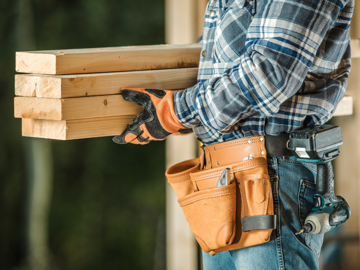 Person in a plaid shirt, wearing work gloves and a tool belt, carries four wooden planks at a construction site—an example you might see in a Restoration Glossary. A cordless drill is attached to their belt.