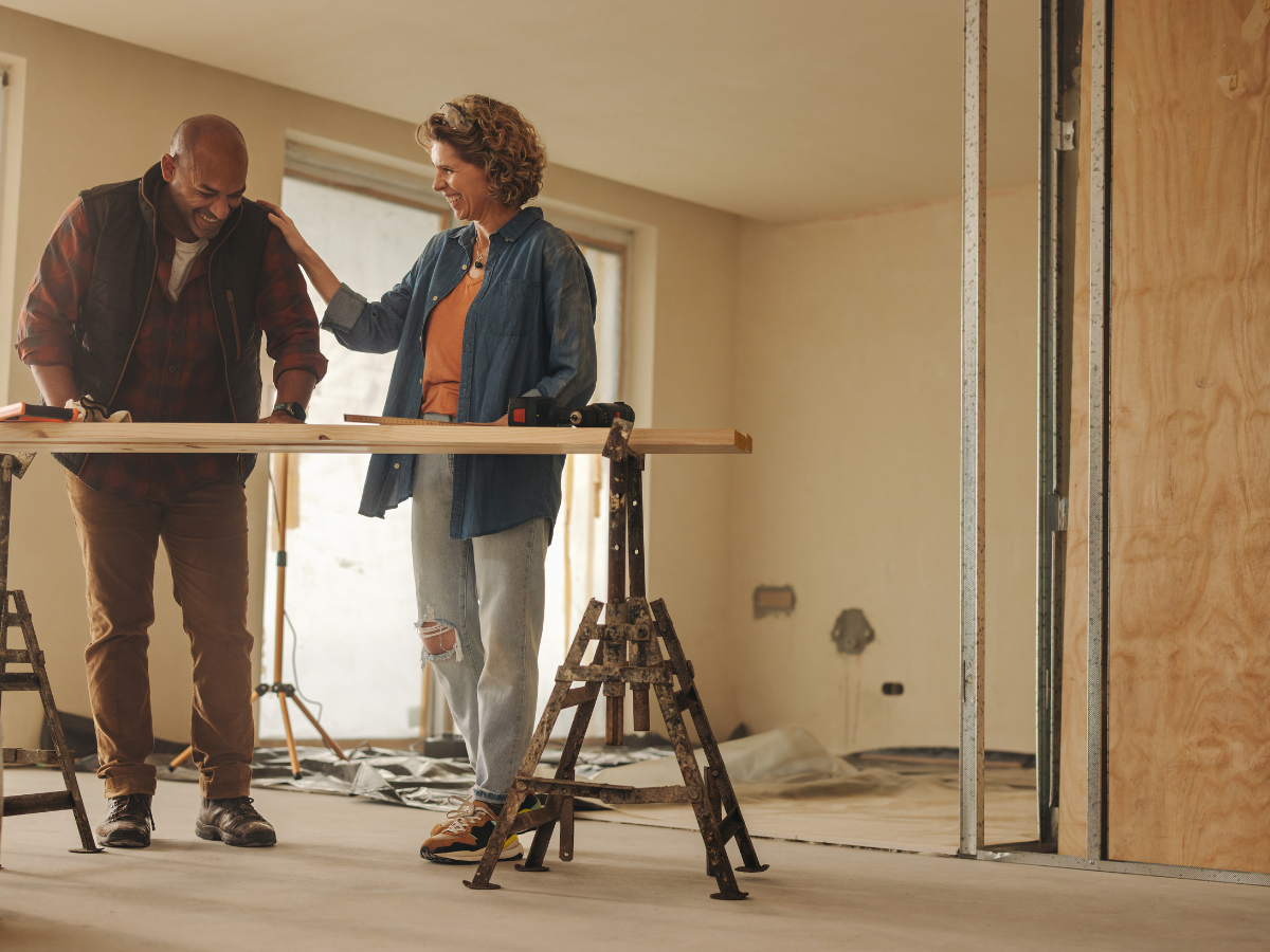 Two people stand in a partially renovated room, smiling and working together on a piece of wood supported by sawhorses, referencing a Restoration Glossary as construction materials fill the background.