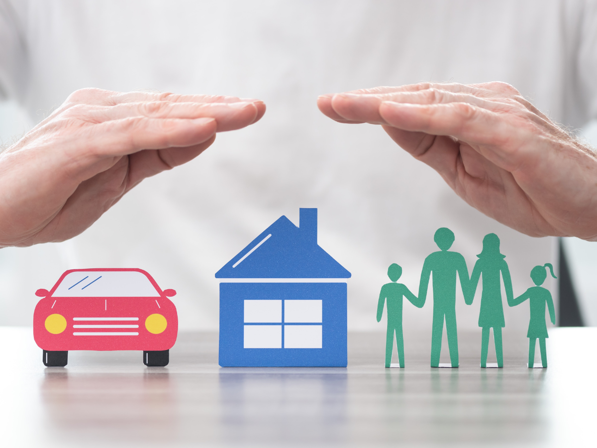 Hands hovering protectively over paper cutouts of a red car, a blue house, and a green family, symbolizing insurance or protection for assets and loved ones—key concepts in any Restoration Glossary.