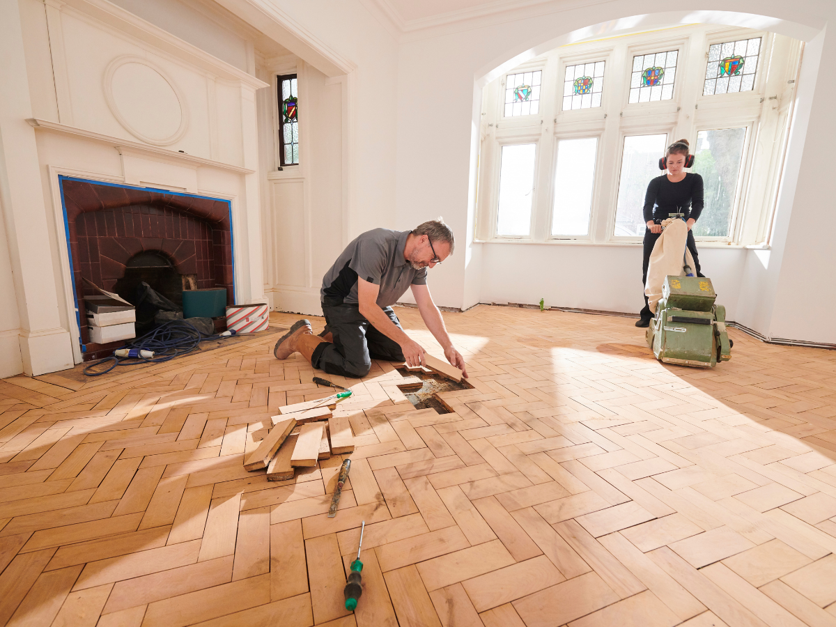Two people working on a herringbone wood floor; one fits planks while the other operates a floor sander, demonstrating techniques often found in the Restoration Glossary, in a sunlit room with large windows.