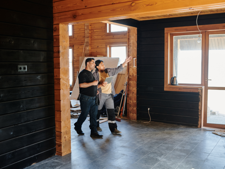 Two people stand in a partially renovated wooden room, one pointing toward the ceiling while discussing the space. Construction materials and a Restoration Glossary are visible in the background.