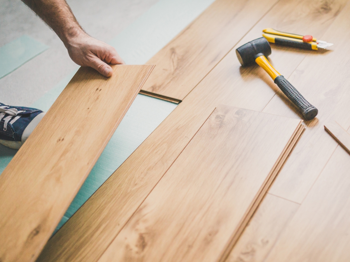 A person installing laminate wood flooring to help fix squeaky floors, with a rubber mallet, utility knife, and ruler placed on top of the panels.