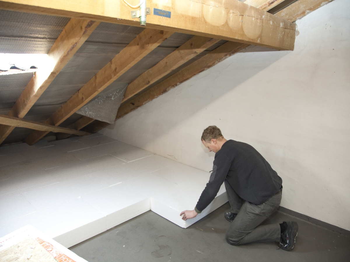 A person kneels on the floor of an attic, installing large white insulation panels along the wall and under the sloped ceiling to help fix squeaky floors and improve overall comfort.