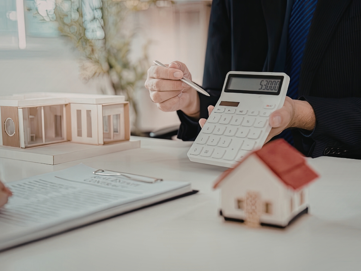 Person in a suit holding a calculator displaying 52,000, with model houses and documents on a desk in an office setting—perhaps planning budgets to fix squeaky floors or manage other home repairs.