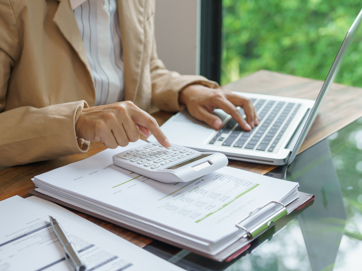 Drywall Vs Plaster: What Homeowners Should Know 3 Person in a tan jacket using a calculator and laptop, working with financial documents on a wooden desk near a window—researching costs for drywall vs plaster.