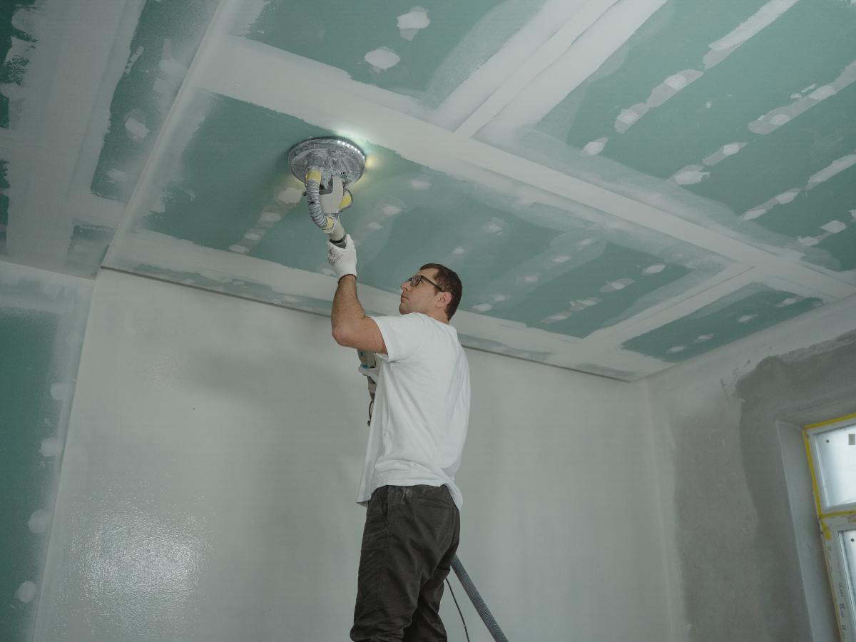 Drywall Vs Plaster: What Homeowners Should Know 4 A man stands on a ladder sanding the ceiling of a room under construction, holding an electric sander with a light—showcasing the process often involved in finishing drywall vs plaster surfaces.