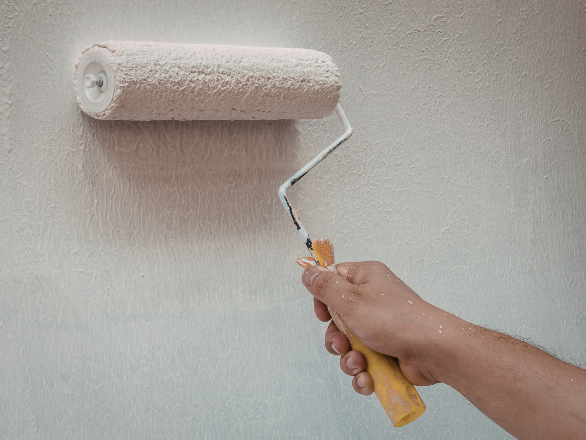 Drywall Vs Plaster: What Homeowners Should Know 6 A hand holding a paint roller applies a fresh coat of white paint to a textured wall, highlighting the subtle differences between drywall vs plaster surfaces.