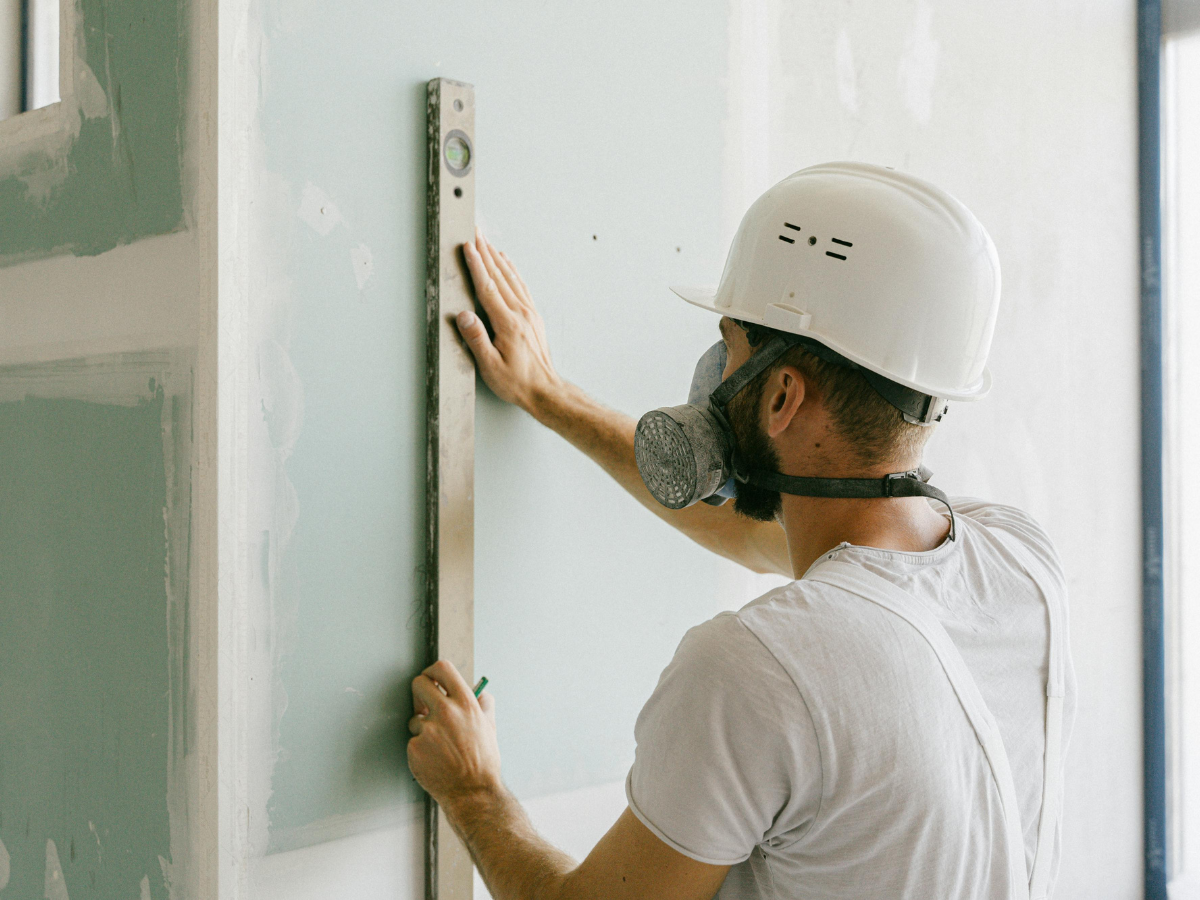 Drywall Vs Plaster: What Homeowners Should Know 7 A construction worker wearing a hard hat and respirator uses a spirit level to measure and align a drywall panel, highlighting precision in the Drywall vs Plaster installation process.