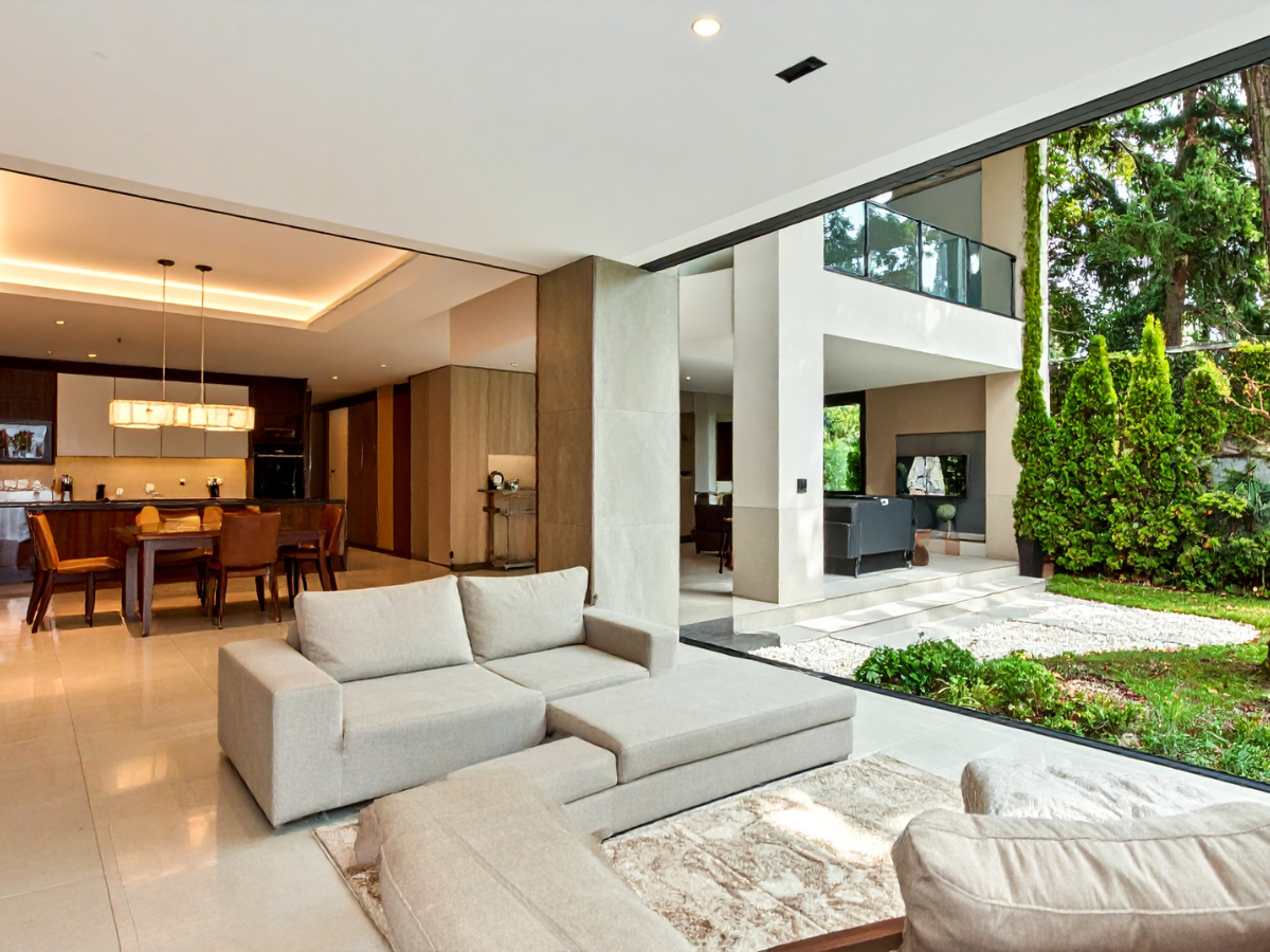 Modern open-concept living room with beige sectional sofas, minimalist design elements, large sliding glass doors, and a view of a landscaped backyard adjacent to a dining area and kitchen.