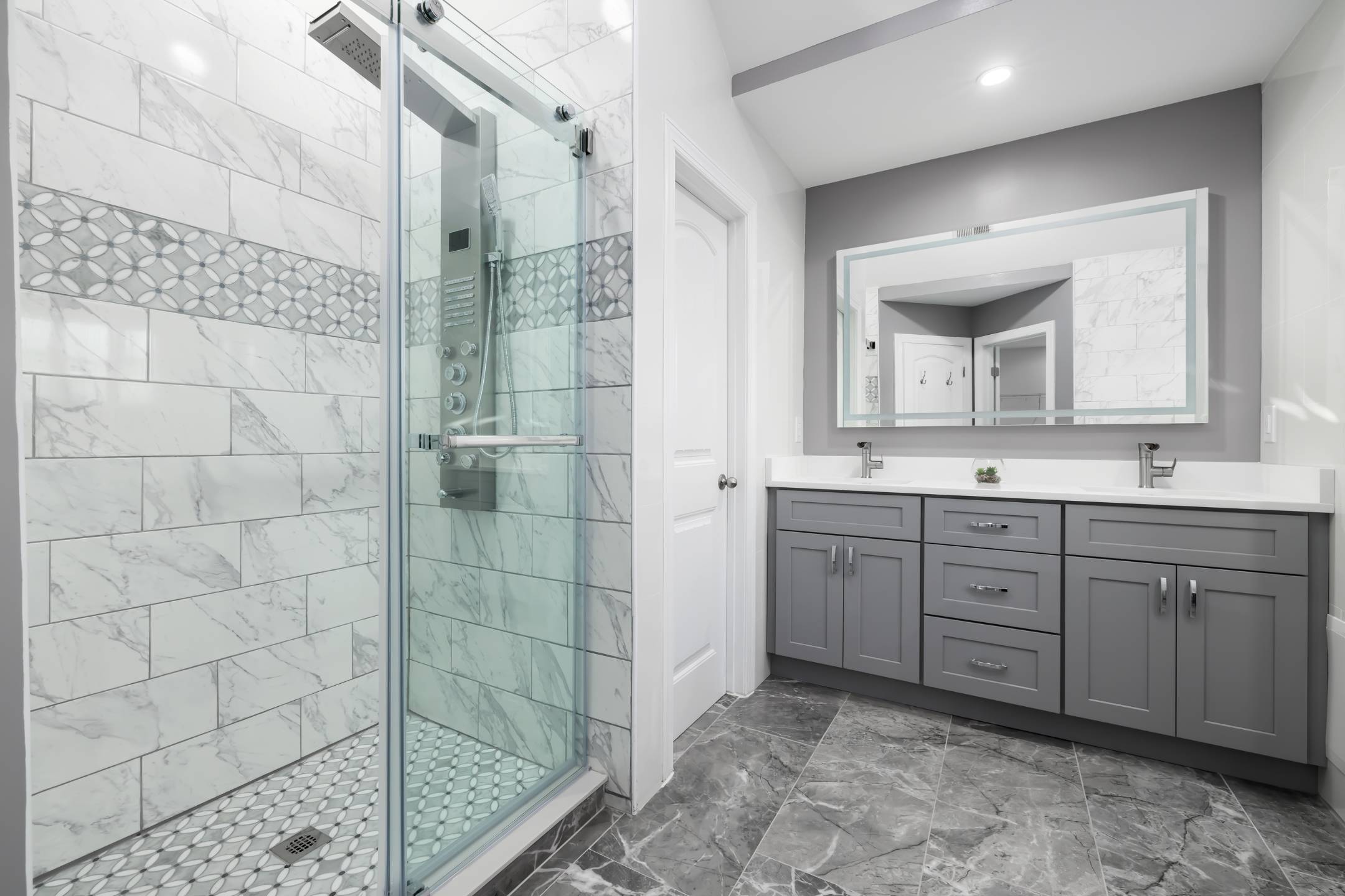 Bathroom And Shower Remodeling 18 Modern bathroom remodeling with a glass-enclosed shower, gray and white tile accents, double-sink vanity with gray cabinets, and marble-patterned flooring.