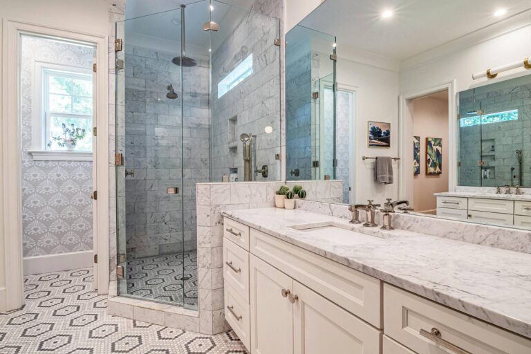 Bathroom And Shower Remodeling 6 Modern bathroom with a glass-enclosed walk-in shower, perfect for shower remodeling, marble countertops, white cabinets, patterned tile floor, and wall-mounted fixtures.