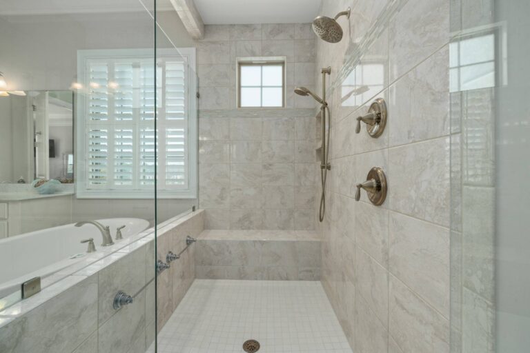 Bathroom And Shower Remodeling 10 Modern tiled walk-in shower with built-in bench, glass enclosure, and two showerheads is a stunning example of shower remodeling, complemented by an adjacent bathtub with window and plantation shutters in the background.