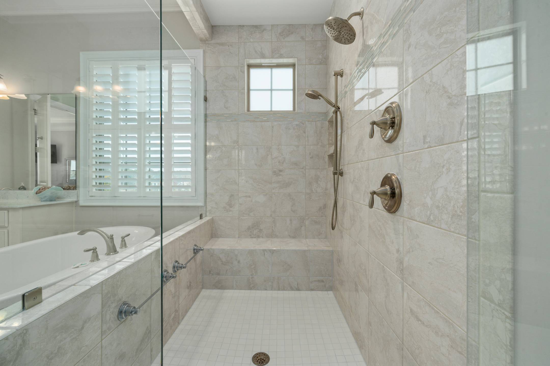 Services 8 Modern tiled walk-in shower with built-in bench, glass enclosure, and two showerheads is a stunning example of shower remodeling, complemented by an adjacent bathtub with window and plantation shutters in the background.