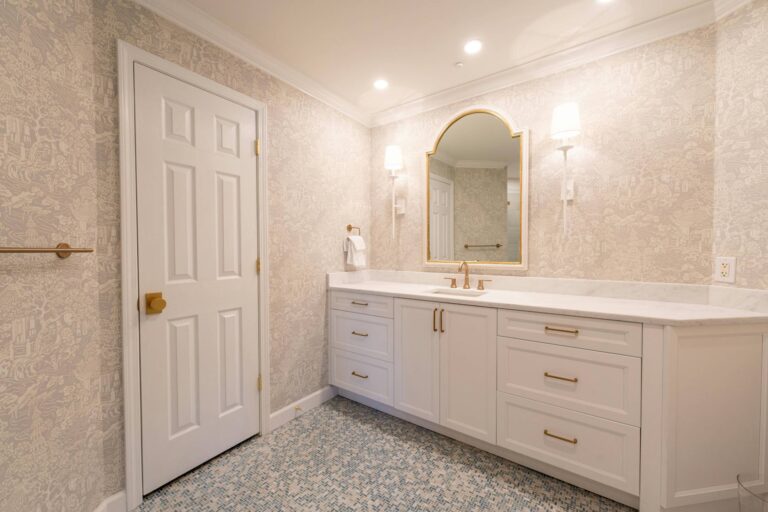 Bathroom And Shower Remodeling 11 Modern bathroom with white cabinets, gold hardware, a large mirror, wall-mounted lights, patterned wallpaper, and a closed door—perfect inspiration for your next bathroom remodeling or shower remodeling project.