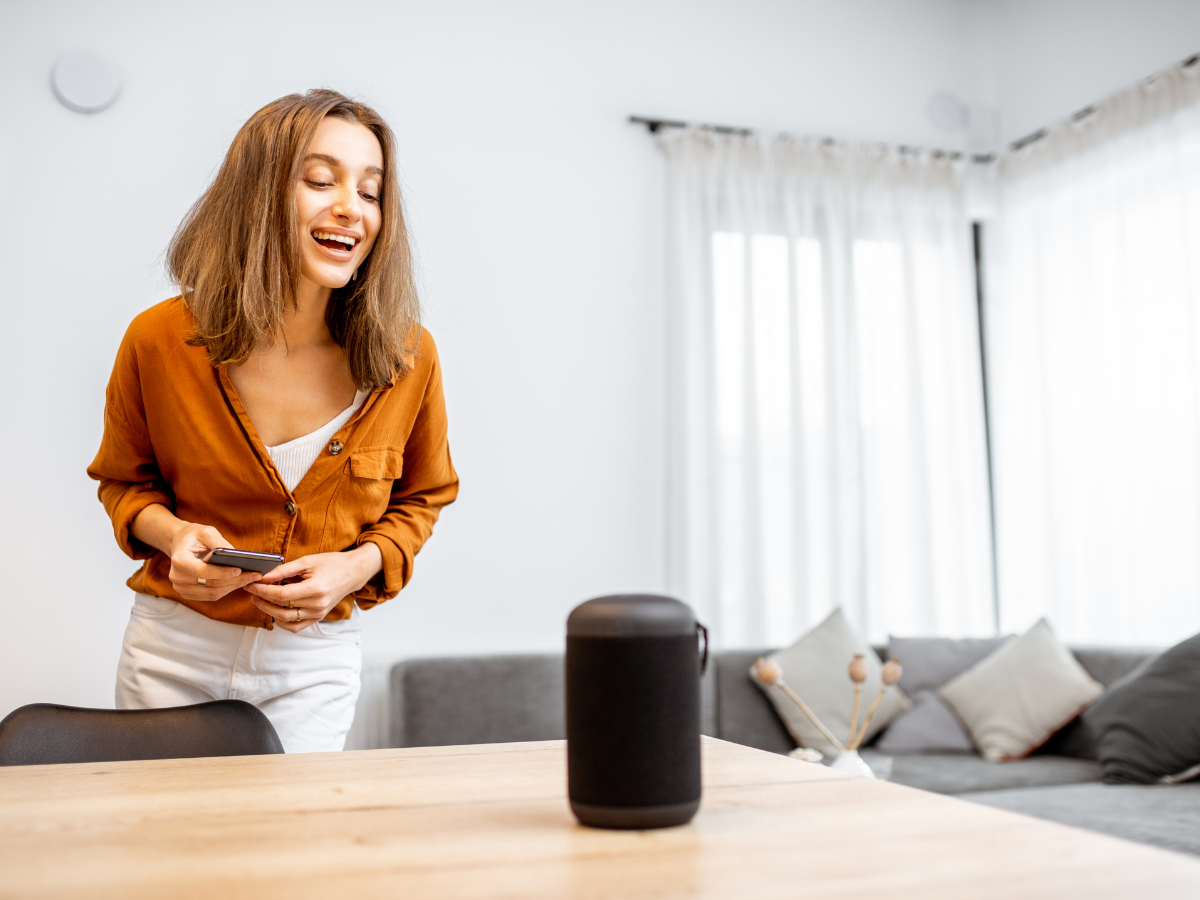 Smart Home Automation For Modern Living 3 A woman stands at a table, smiling while interacting with a smart speaker and holding a smartphone, enjoying seamless home automation in a bright living room.