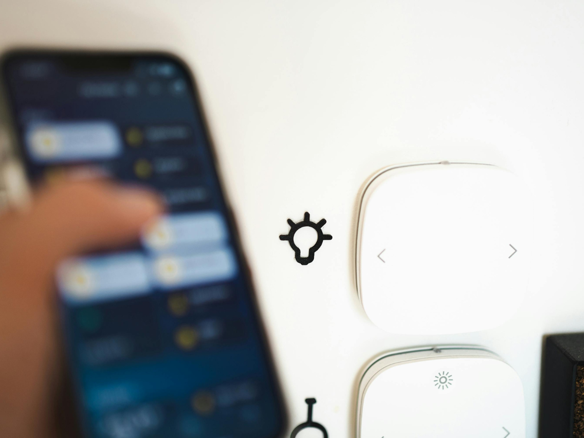 Smart Home Automation For Modern Living 4 A person uses a smartphone in front of a wall-mounted home automation control panel with two white devices and a lightbulb icon.