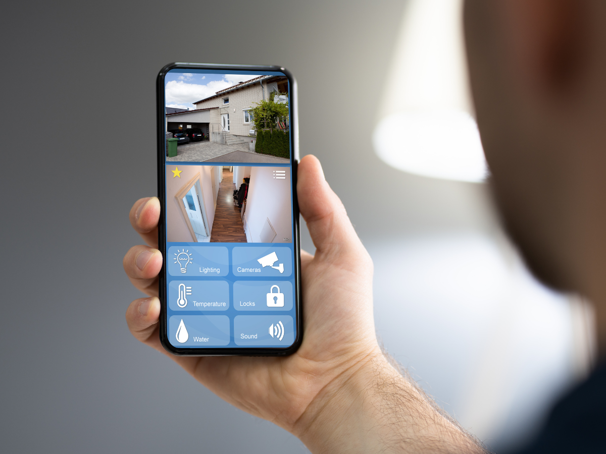 Smart Home Automation For Modern Living 5 Person holding a smartphone displaying a home security app with live camera feeds and home automation features for controlling lighting, sound, temperature, locks, and water.