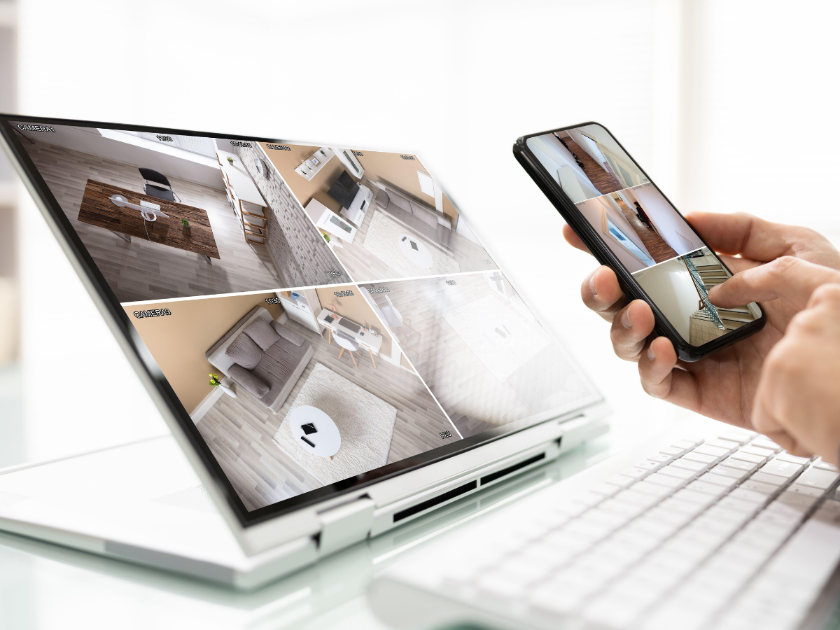 Smart Home Automation For Modern Living 7 A person monitors security camera footage showing different rooms on both a laptop and a smartphone beside a computer keyboard, highlighting the convenience of home automation.