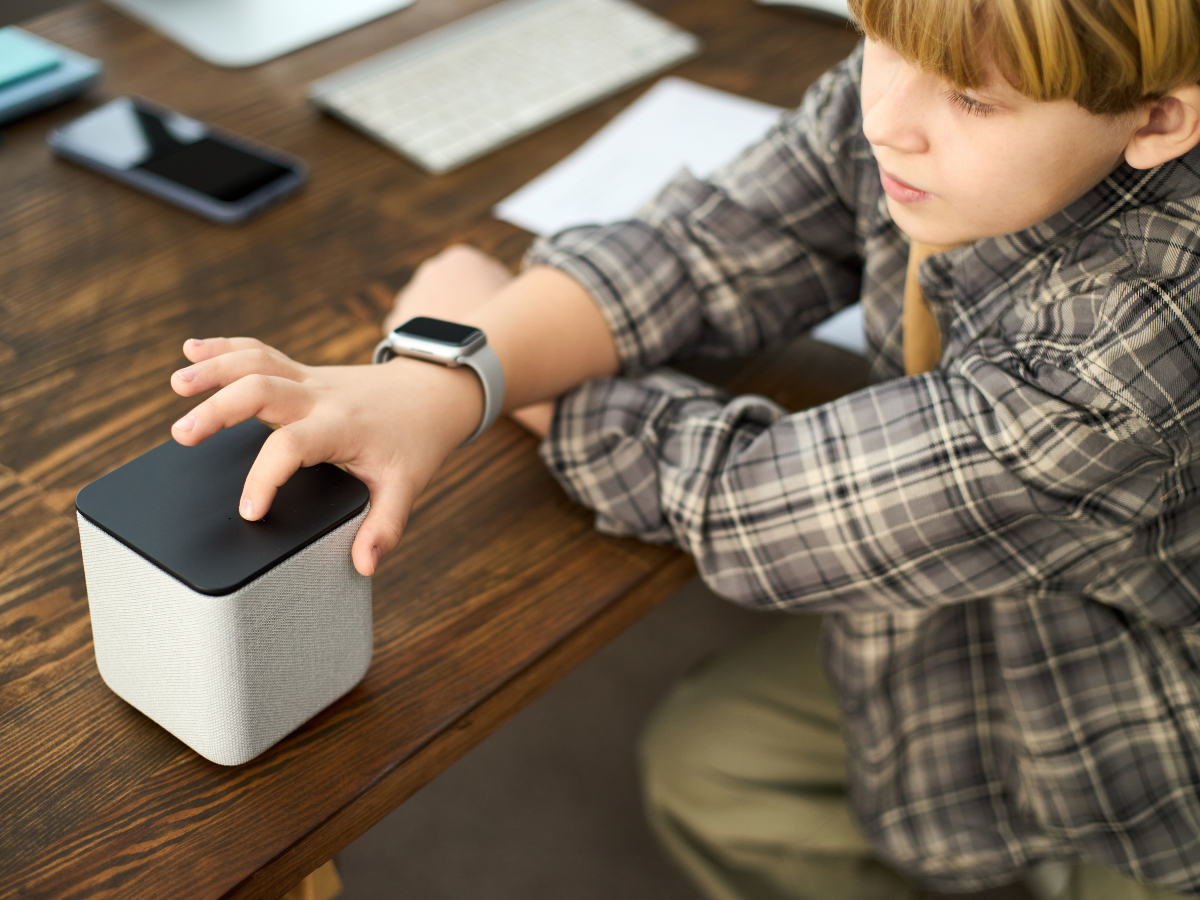 Smart Home Automation For Modern Living 8 A child in a plaid shirt touches the top of a smart speaker on a wooden desk, showcasing home automation, with a smartwatch on their wrist and electronics in the background.