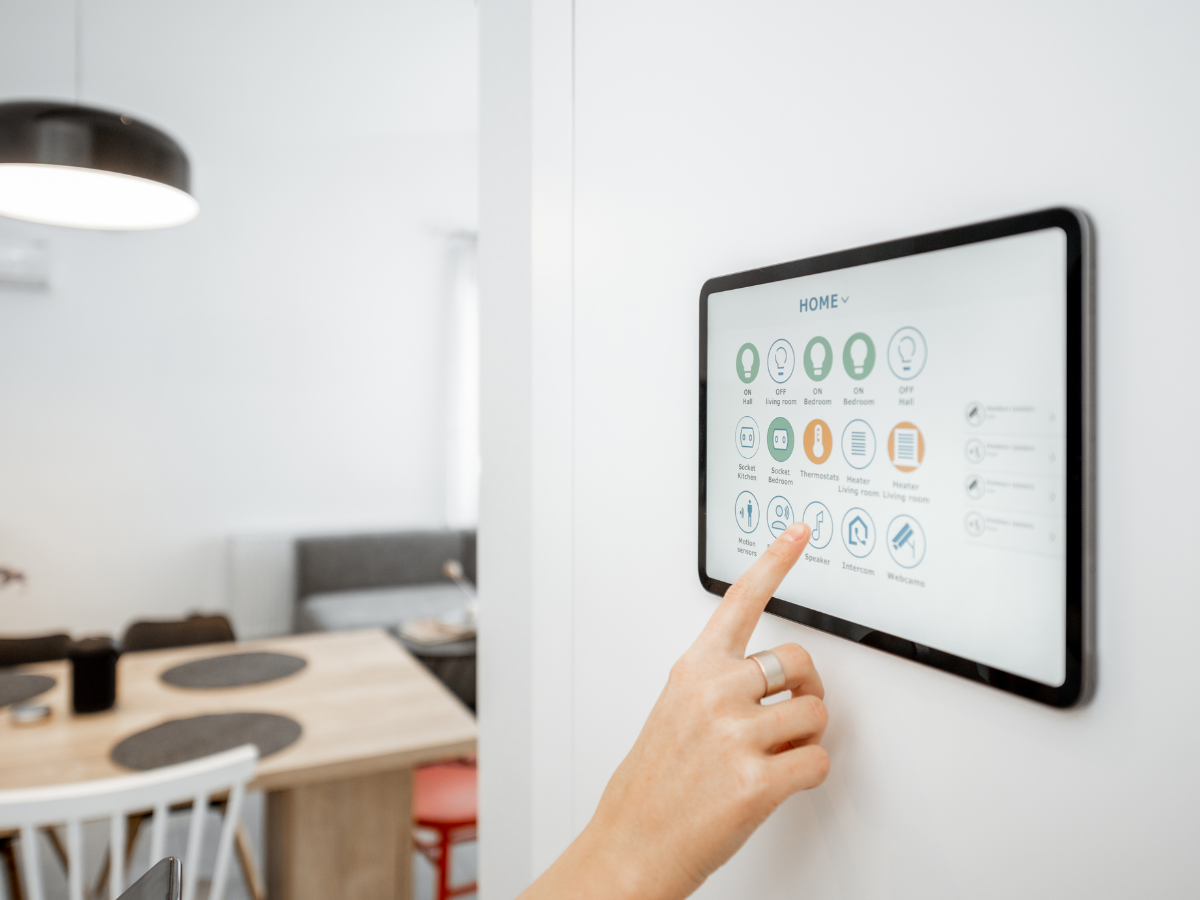 Smart Home Automation For Modern Living 1 A person uses a wall-mounted touchscreen control panel for home automation functions in a modern kitchen and dining area.