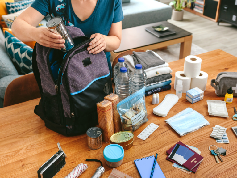 A person packs a backpack with canned food, water bottles, toiletries, medicine, clothes, and emergency supplies on a wooden table for storm prep.