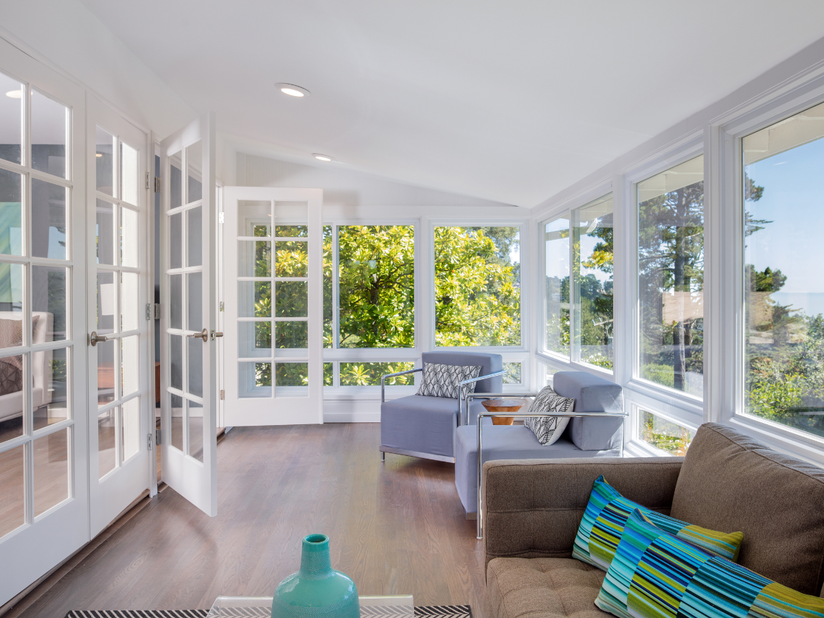 Bright sunroom with large windows, white walls, wooden floor, a gray armchair, brown sofa, accent pillows, and a turquoise vase on a table&mdash;expertly crafted by sunroom builders Atlanta. Trees and sky visible outside.