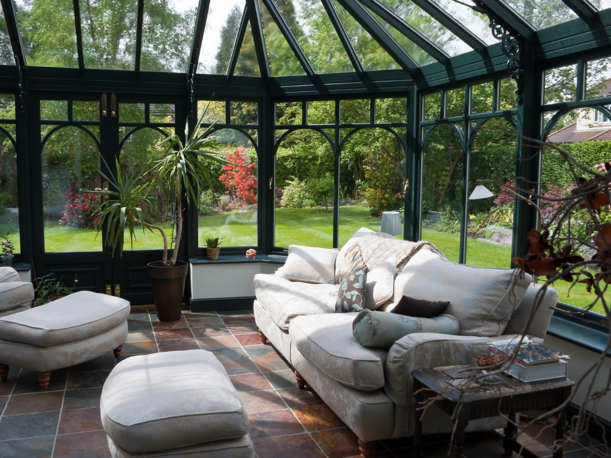 A sunlit conservatory, designed by expert sunroom builders Atlanta, features green-framed glass walls, a comfortable sofa, armchairs, potted plants, and elegant tiled flooring&mdash;all overlooking a lush garden.