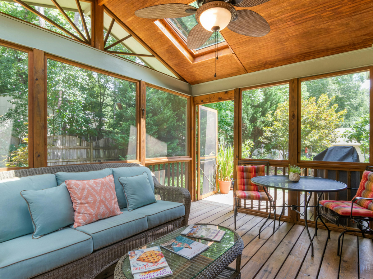 A sunroom with large screened windows, expertly crafted by sunroom builders Atlanta, features wicker furniture with colorful cushions, a round table with chairs, and a ceiling fan overhead.