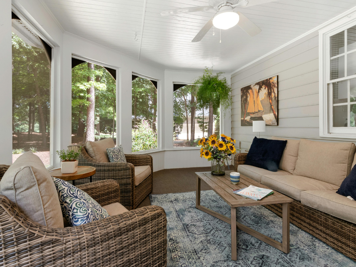 A sunroom with wicker furniture, tan cushions, a coffee table with sunflowers, a hanging fern, and large windows overlooking trees&mdash;expertly crafted by sunroom builders Atlanta to create a peaceful retreat filled with natural light and scenic views.
