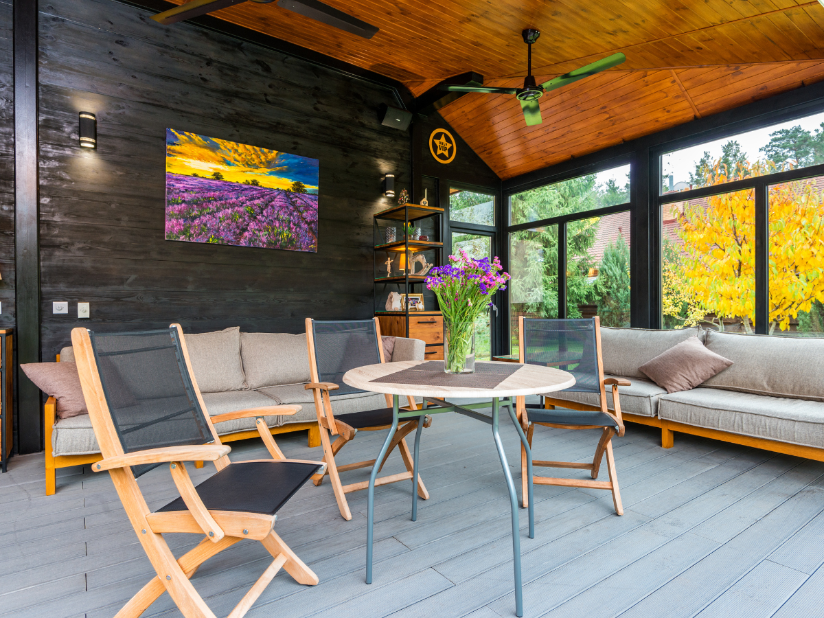 Modern sunroom with black walls, wooden ceiling, large windows, beige sofas, wooden chairs, a round table with flowers, and a colorful landscape painting on the wall&mdash;a stunning example of the craftsmanship offered by top sunroom builders Atlanta.