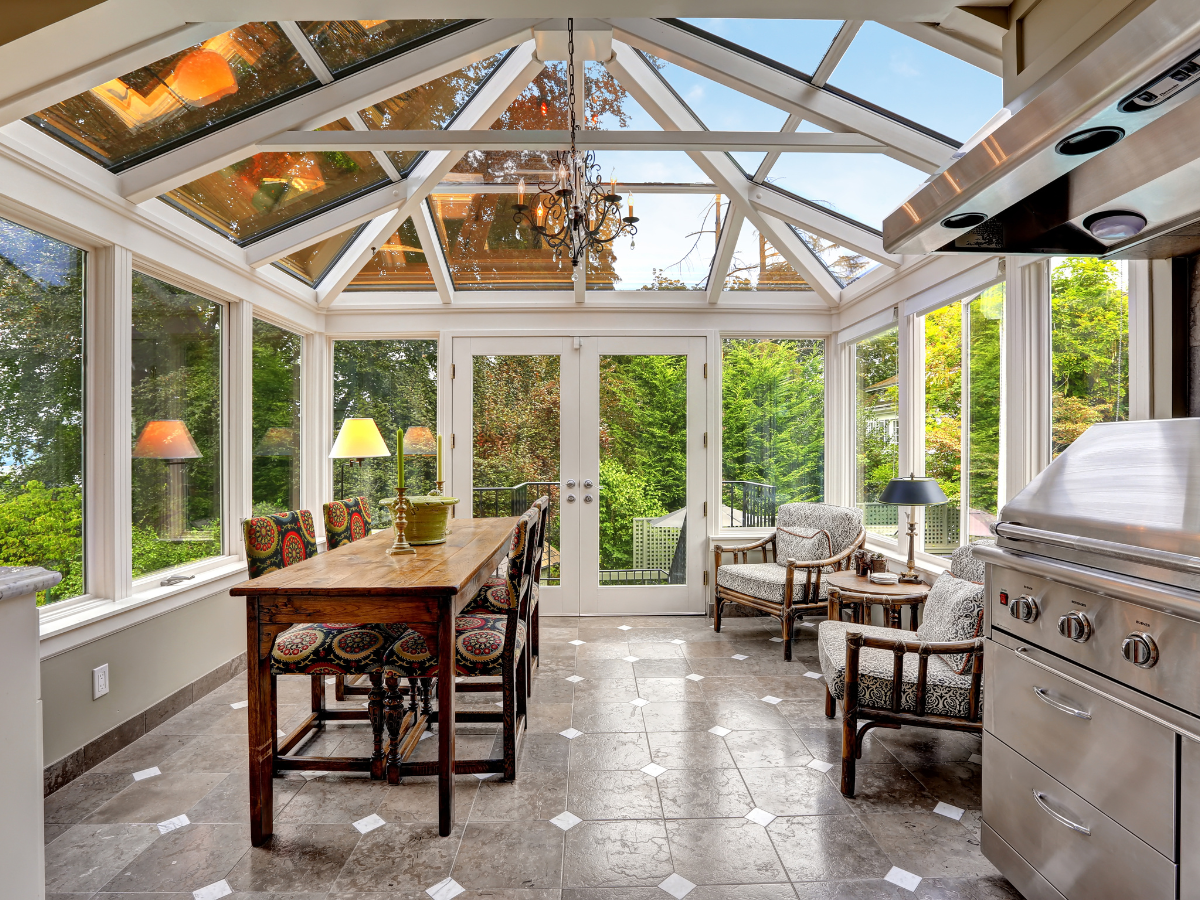 Bright sunroom with large windows and glass ceiling, expertly crafted by premier sunroom builders in Atlanta. This inviting space features a wooden dining table, four patterned chairs, two armchairs, and a stainless steel grill, all overlooking a lush green outdoor view.