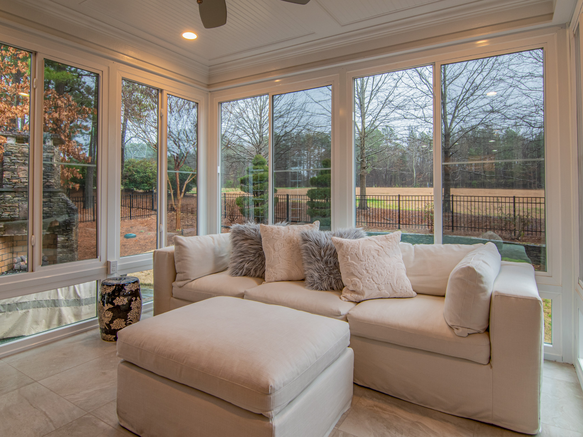 A bright sunroom with large windows, a white sectional sofa with throw pillows, and a view of a yard with trees and a black fence showcases the expert craftsmanship of sunroom builders Atlanta.