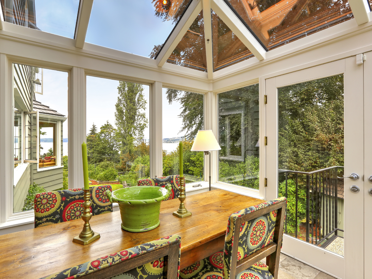 Sunroom builders Atlanta have truly crafted a beautiful space featuring a wooden table, patterned chairs, a green centerpiece, two brass candlesticks, and a lamp. Large windows highlight the trees and water outside, making this sunroom an inviting retreat.