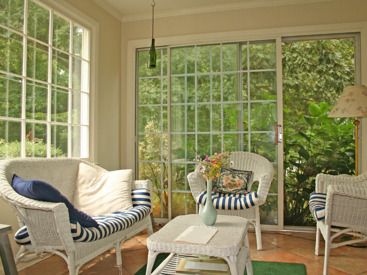 Sunroom builders Atlanta have crafted this inviting sunroom with white wicker furniture, blue-striped cushions, a small table with a vase of flowers, and large windows overlooking lush greenery.