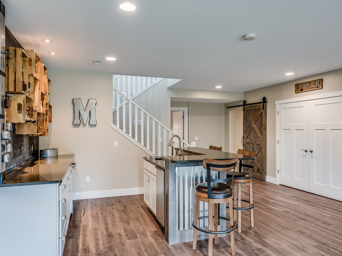 Modern basement with wood flooring, a metal bar, three barstools, stairs, a barn door, and wall d&eacute;cor including a large letter &ldquo;M&rdquo; and a &ldquo;River&rdquo; sign&mdash;an inspiring example of basement remodeling Atlanta homeowners will love.