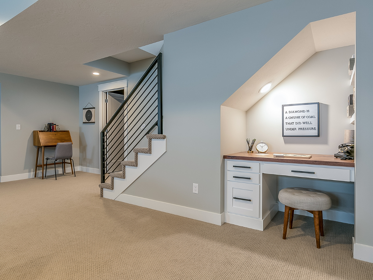 A basement room with a carpeted floor, a staircase featuring a metal railing, a built-in desk tucked under the stairs, and a small writing desk against the wall&mdash;this inviting space showcases basement remodeling Atlanta style at its finest.