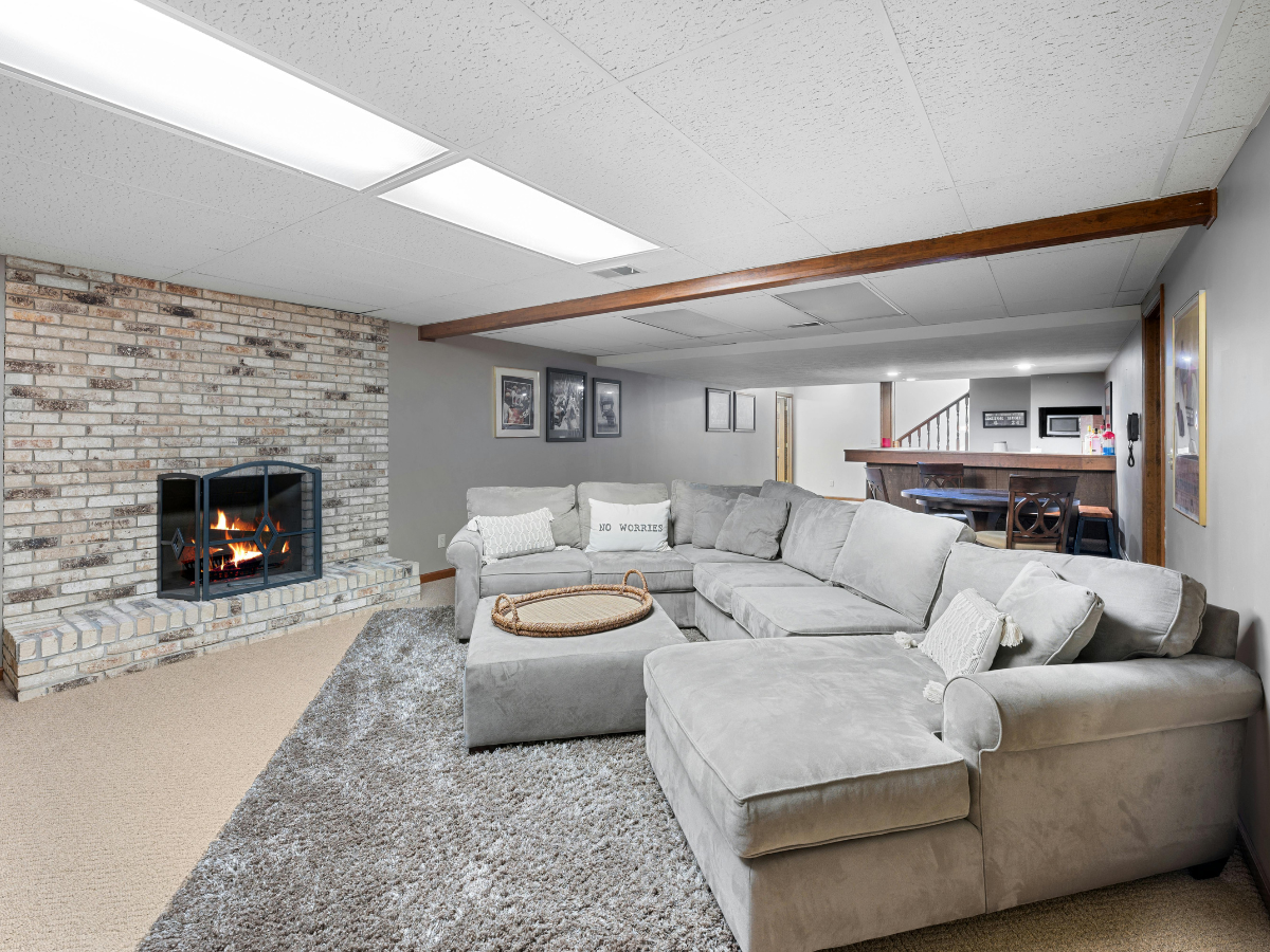Finished basement with gray sectional sofa, brick fireplace, gray carpet, and wood beam ceiling; framed pictures and a desk area in the background—perfect inspiration for those considering basement remodeling Atlanta projects.