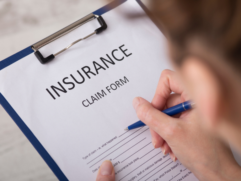 Person filling out an insurance claim form on a clipboard with a blue pen; the form has blank spaces for personal and claim details, suggesting the need for insurance claim assistance.