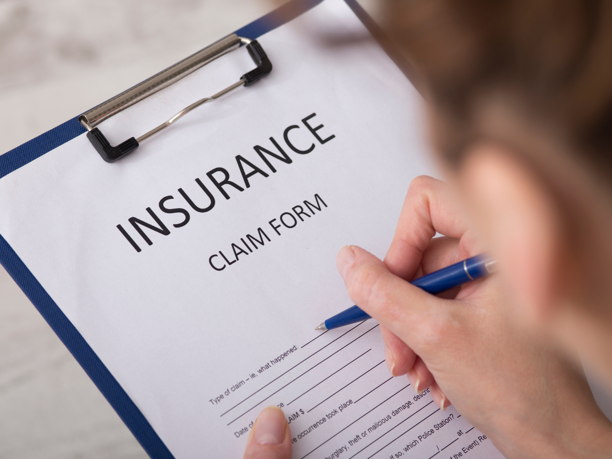 Person filling out an insurance claim form on a clipboard with a blue pen; the form has blank spaces for personal and claim details, suggesting the need for insurance claim assistance.
